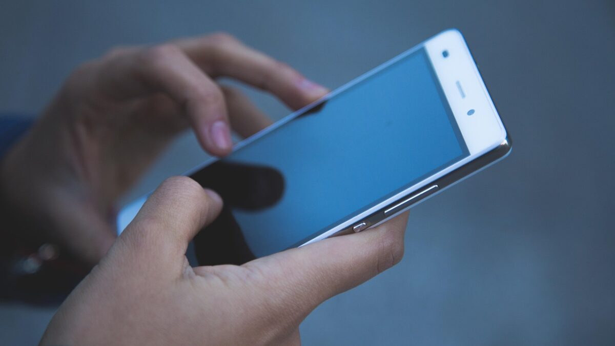 Gros plan sur deux mains tenant un smartphone blanc à écran bleuté, l'une pointant l'écran, sur fond flou bleu-gris.