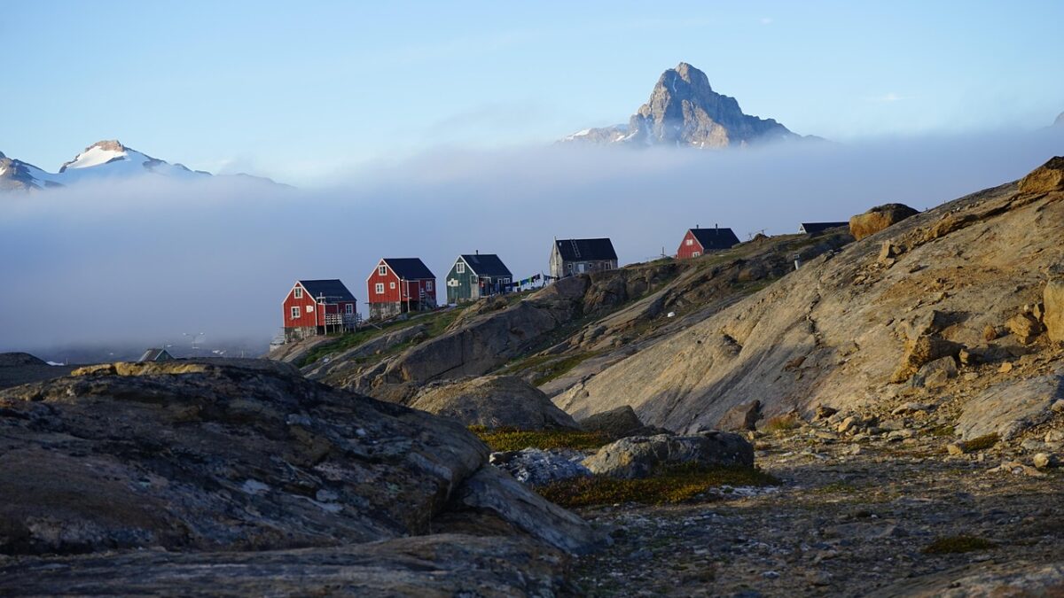 Village groenlandais au bord d'une côte rocheuse, avec plusieurs maisons en bois rouges, vertes et grises sur une colline, une brume basse et des montagnes enneigées sous un ciel bleu.