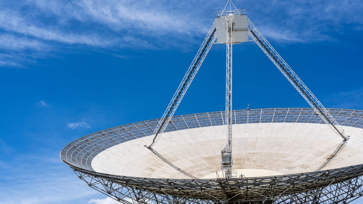 Grand radiotélescope parabolique blanc vu en contre-plongée, avec sa structure métallique en treillis et son mât central, sous un ciel bleu parsemé de quelques nuages.