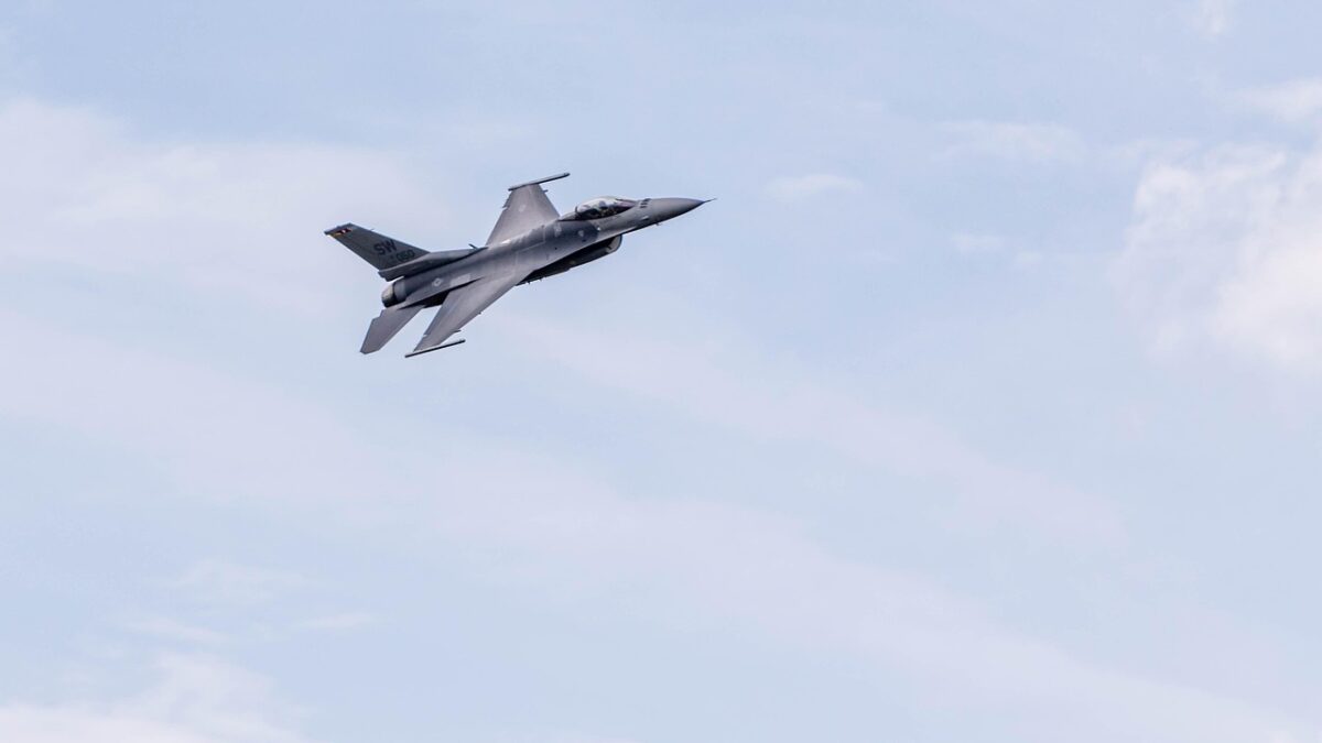 Un avion de chasse gris F-16 en vol dans un ciel bleu nuageux, vu de profil, avec sa verrière visible et des marquages discrets sur la dérive.