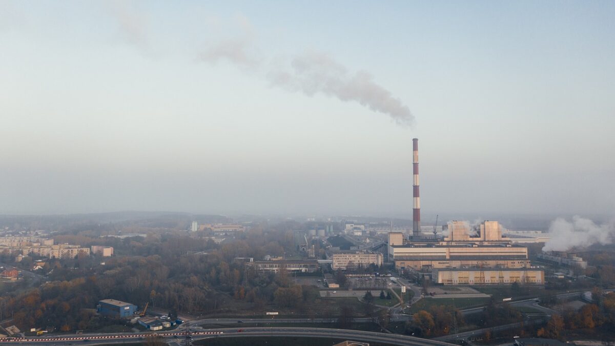 Luftaufnahme einer Industrieanlage mit einem hohen, rot-weiß gestreiften Schornstein, aus dem eine Rauchfahne aufsteigt, umgeben von Gebäuden, Wohngebieten und einer Autobahn im Morgennebel.