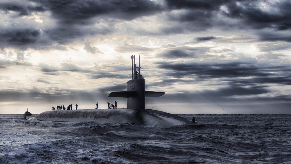 Un sous-marin à propulsion nucléaire en surface, sur une mer agitée sous un ciel nuageux ; plusieurs marins sur le pont, et un petit bateau d'escorte à gauche.