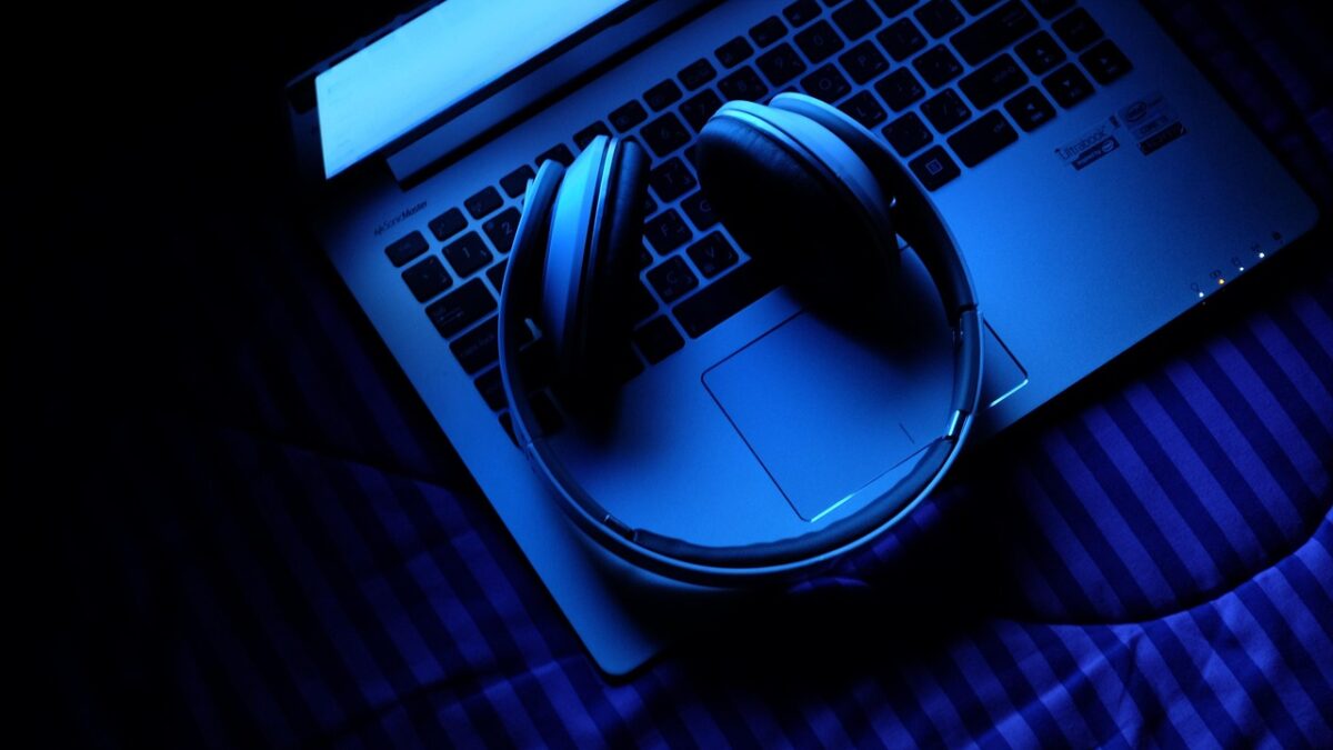 Un ordinateur portable posé sur un lit à rayures bleues, un casque audio posé sur le clavier, dans une ambiance bleue sombre.