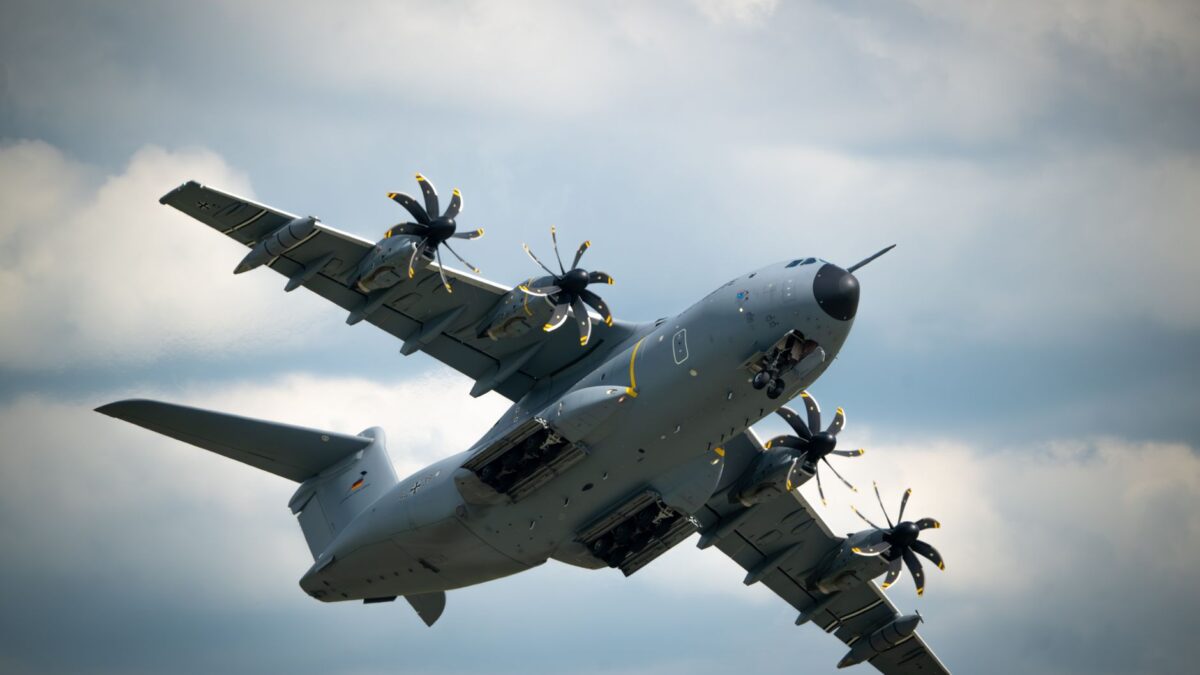 Ein graues militärisches Transportflugzeug (Airbus A400M) fliegt, von unten und seitlich fotografiert, mit vier Propellern mit gelb-schwarzen Blättern vor einem bewölkten Himmel.