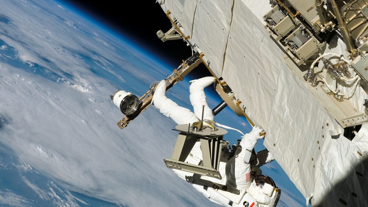 Un astronaute en combinaison blanche effectue une sortie extravéhiculaire, accroché à une structure de la Station spatiale internationale, avec la Terre et son atmosphère bleutée en arrière-plan.
