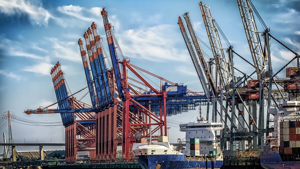 Terminal à conteneurs portuaire avec portiques de quai rouges, bleus et gris, navires de charge et conteneurs empilés sous un ciel bleu nuageux, eau au premier plan.