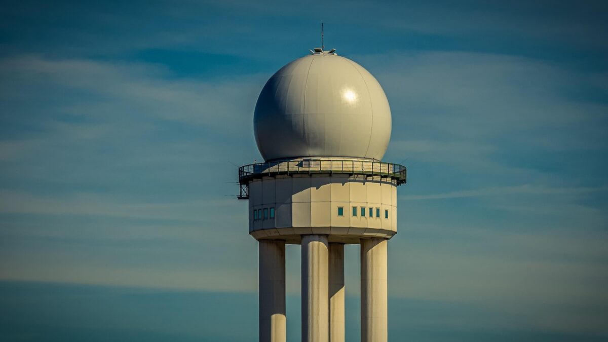 Tour de contrôle d'aéroport avec un radôme blanc sphérique au sommet, sous un ciel bleu parsemé de nuages.