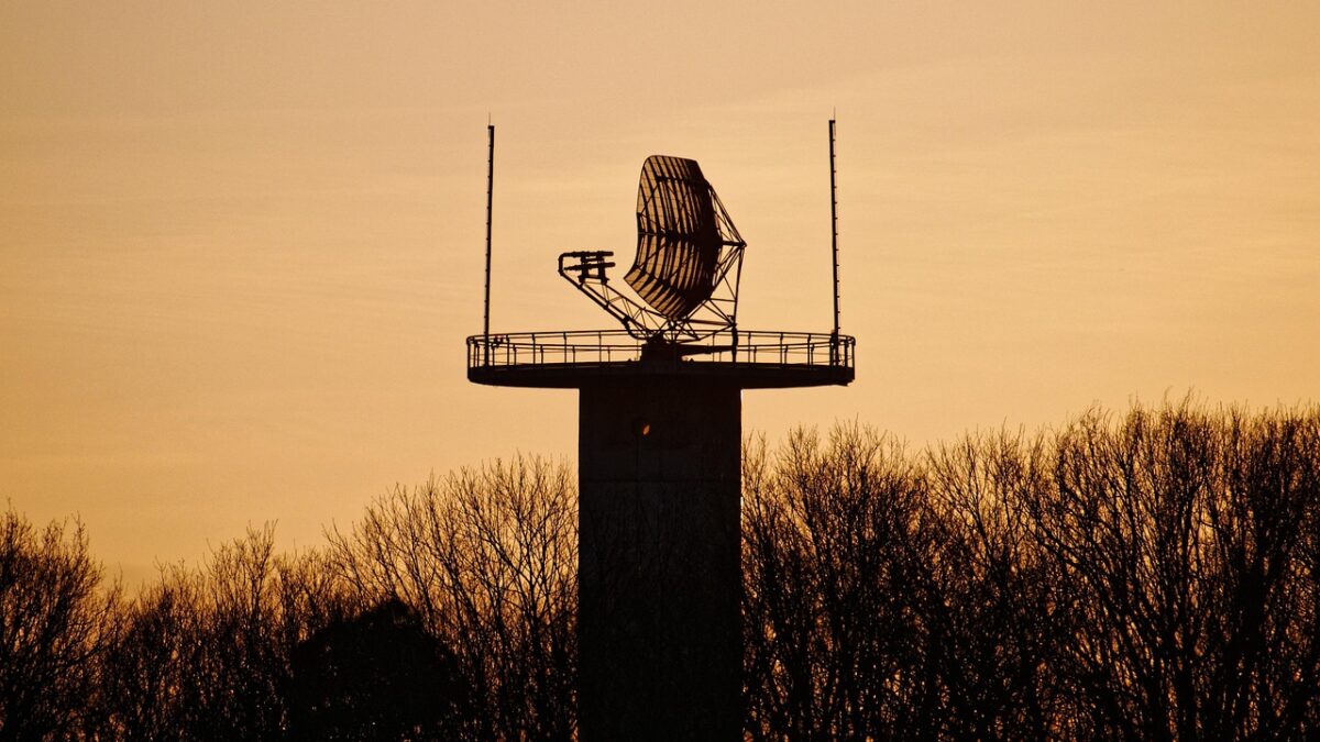 Silhouette d'une antenne radar à réflecteur sur une tour en béton, entourée d'arbres dépouillés, sur fond de ciel orangé au coucher du soleil.