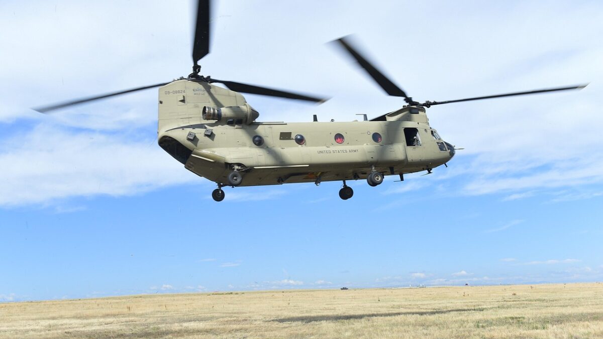 Hélicoptère militaire à double rotor CH-47 Chinook, marqué « UNITED STATES ARMY », en vol stationnaire au-dessus d'une prairie sèche sous un ciel bleu nuageux.