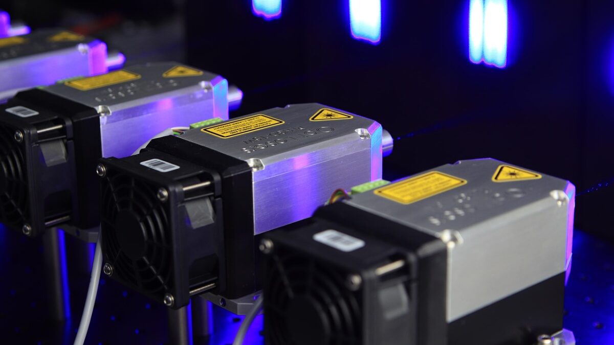 Mehrere schwarze und silberne Metall-Lasermodule mit gelben Warnetiketten liegen auf einem optischen Tisch, blau und violett beleuchtet in einem dunklen Labor.