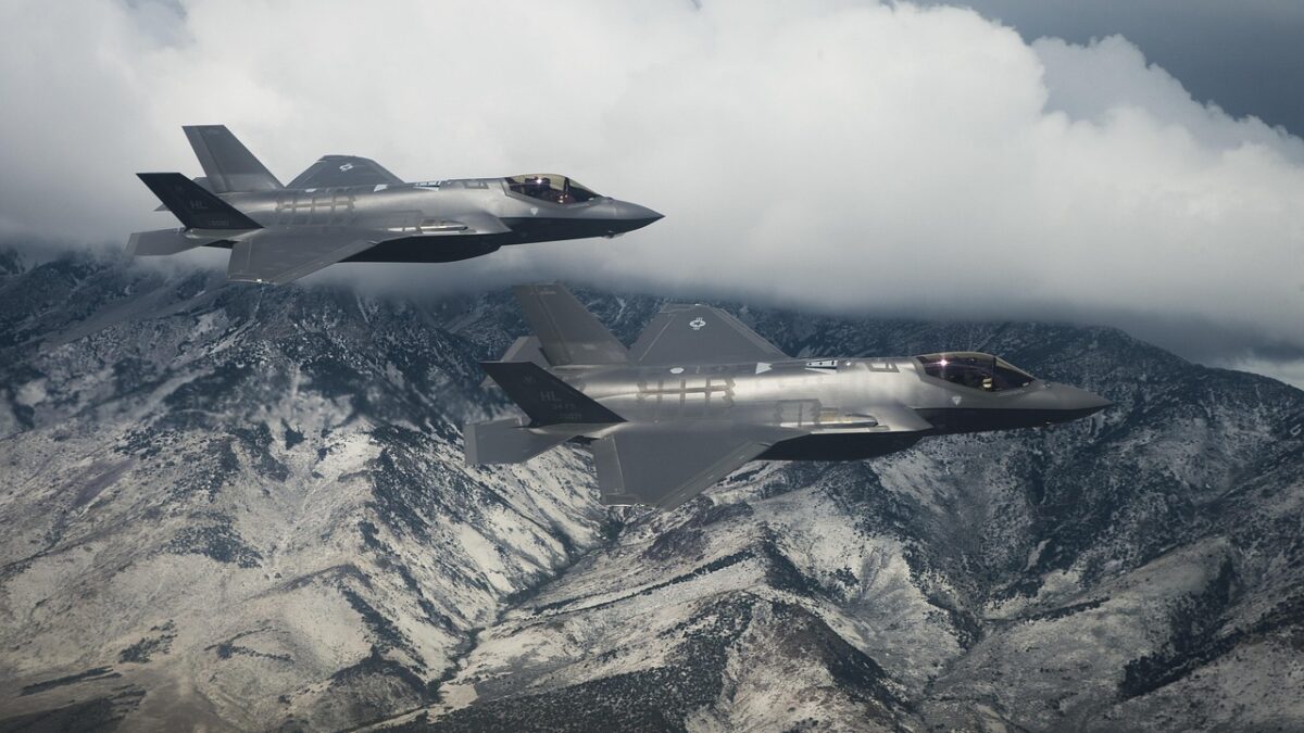Deux avions de chasse furtifs gris volent en formation au-dessus de montagnes enneigées, sous un ciel nuageux.