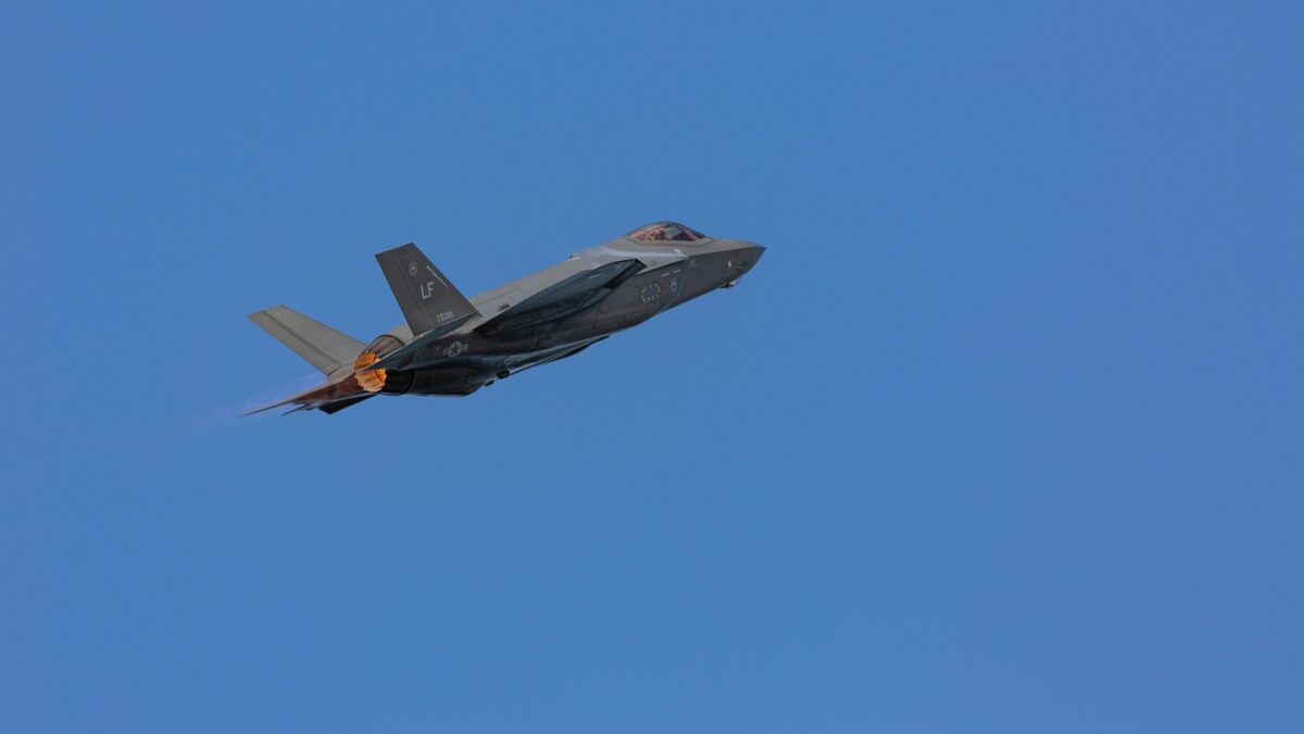 Un avion de combat gris F-35 en vol sur fond de ciel bleu uni, avec une tuyère allumée et la queue marquée « LF ».