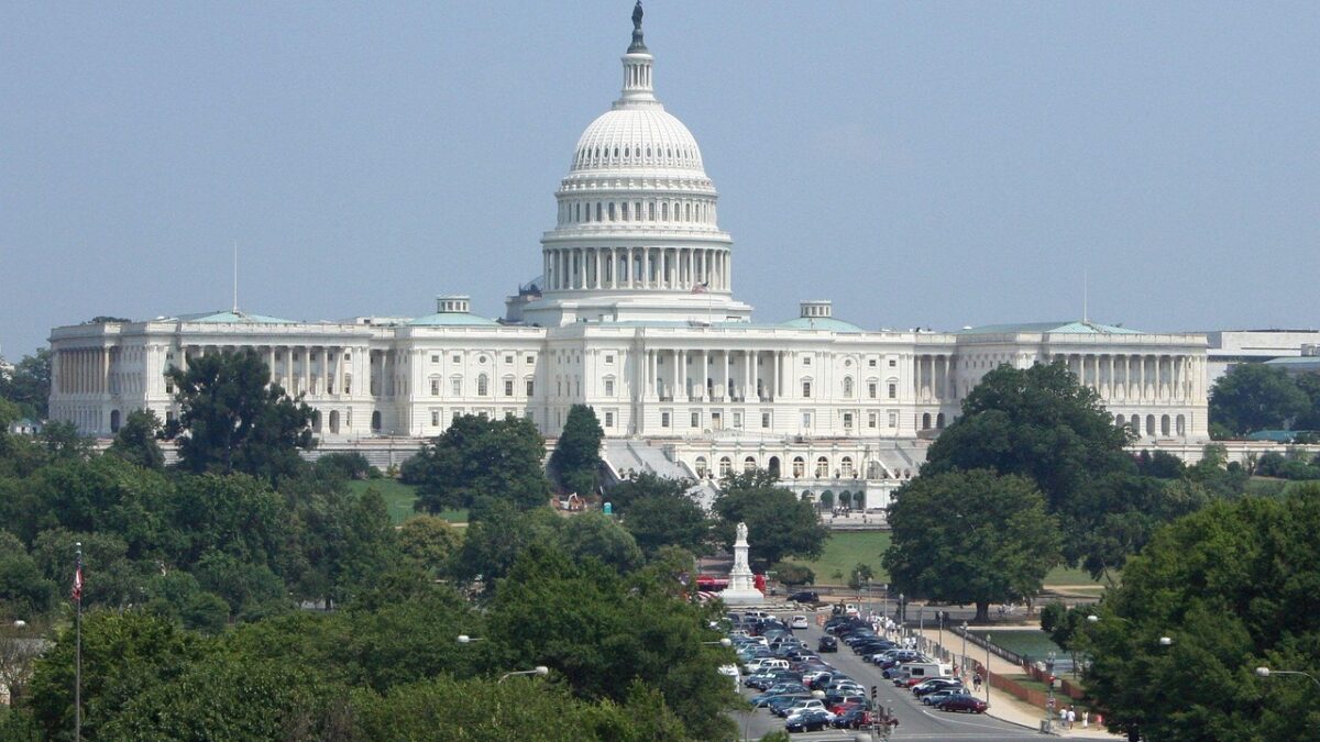 Vue lointaine du Capitole des États-Unis à Washington, avec son dôme blanc, une avenue encombrée de voitures, des arbres et un drapeau américain à gauche.
