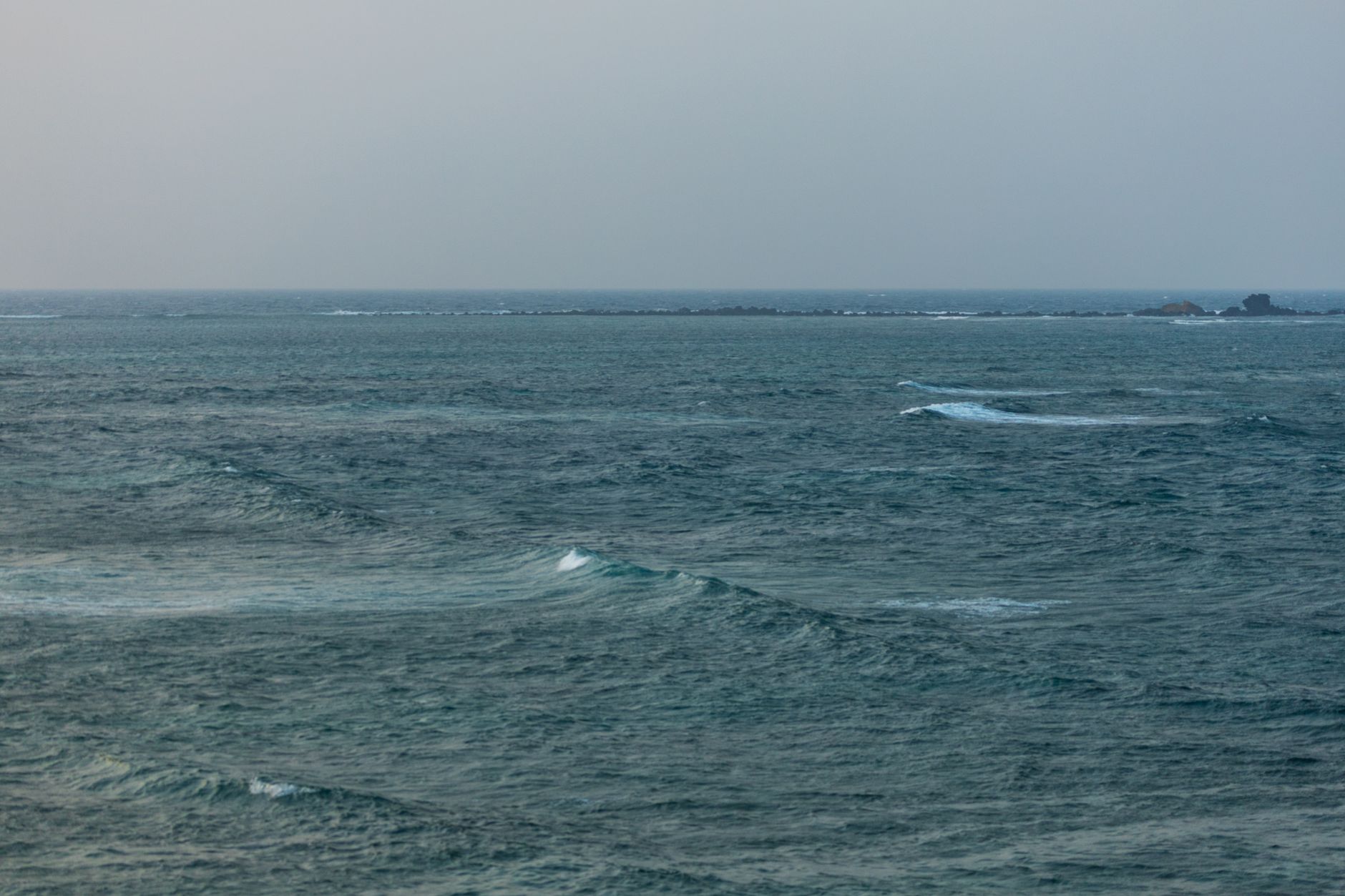 Choppy blue-green sea with small breaking waves, a low rocky islet on the horizon and a hazy grey sky.