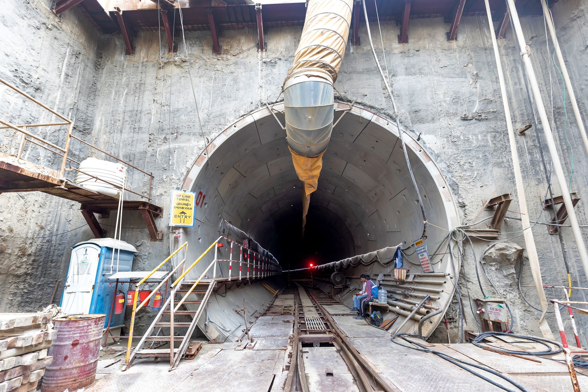 Circular tunnel entrance in a deep excavation with rail tracks, a ventilation duct, cables and scaffolding, and two workers seated near the opening.