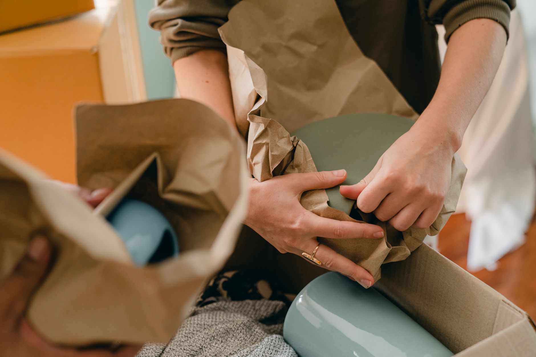 Hands wrapping pale blue-green ceramic items in brown kraft paper and packing them into a cardboard box.