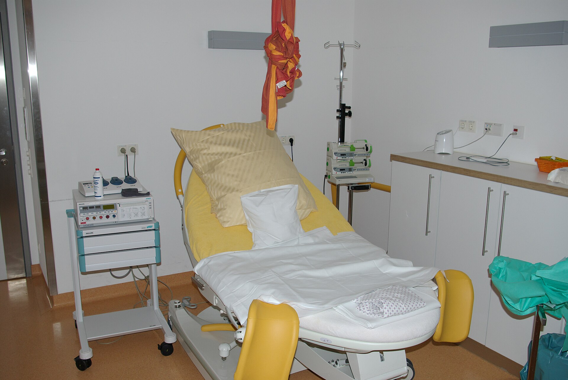 Hospital delivery bed with yellow leg supports, white sheets and pillows, next to a fetal monitor and an infusion stand.