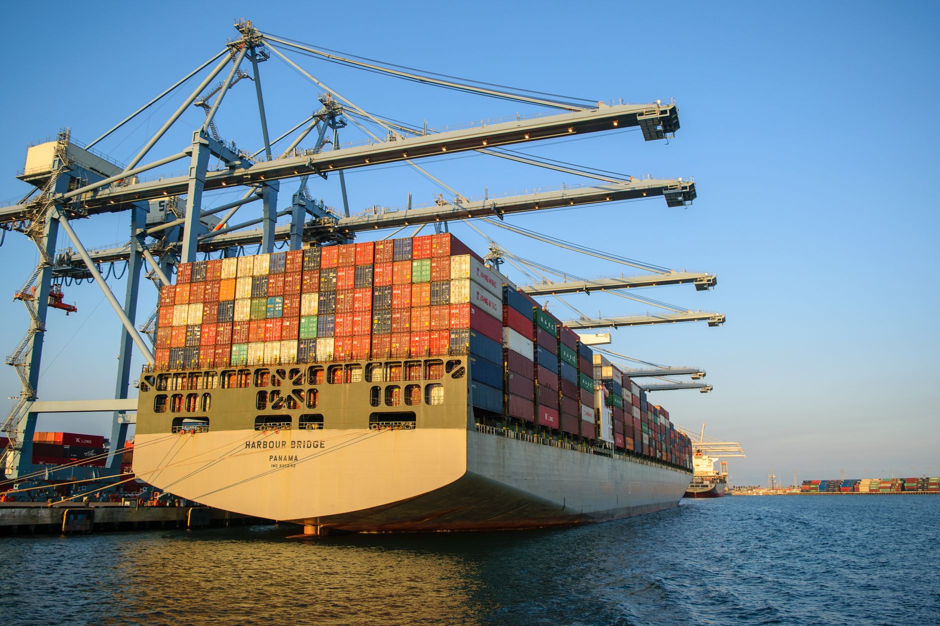 A large container ship loaded with colourful cargo containers docked at a port under tall gantry cranes.