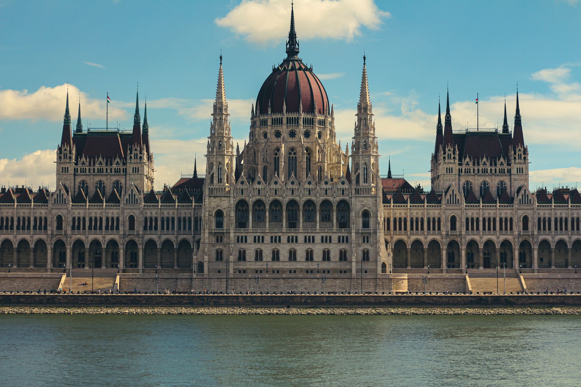 Large Neo-Gothic building with a red central dome and spires, seen across a river under a partly cloudy blue sky.