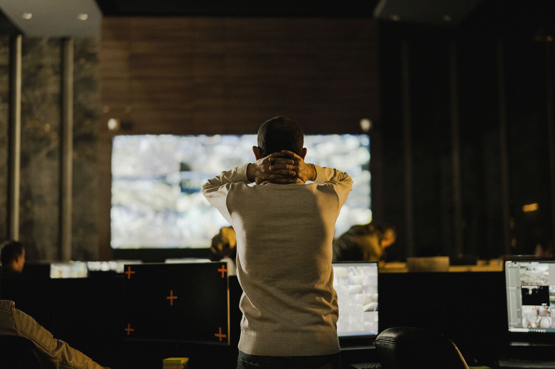 A man seen from behind, hands behind his head, stands in a dark control room facing a large wall screen and several monitors.