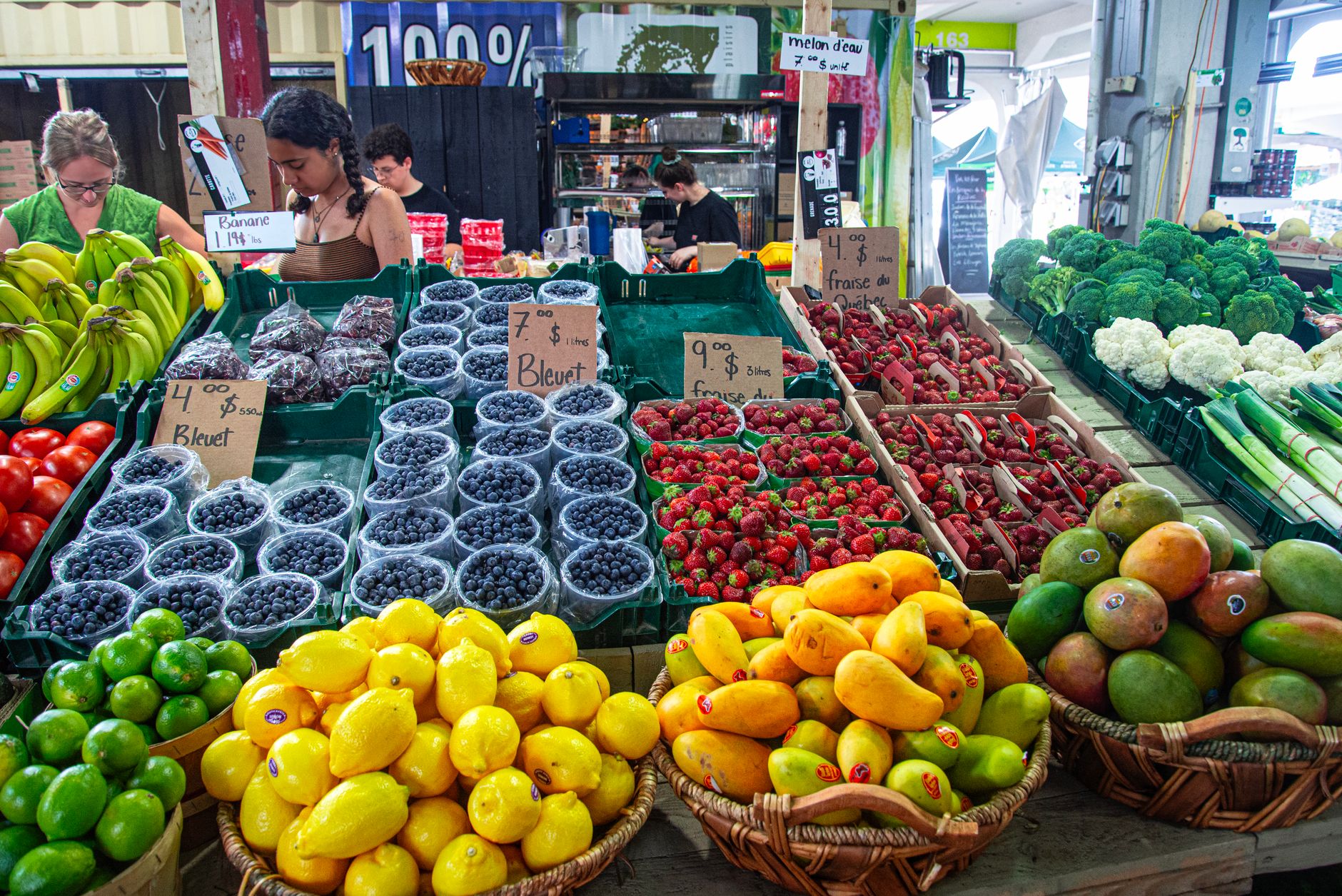 Market stall with blueberries, strawberries, lemons, limes, mangoes, bananas and cauliflower, with vendors and handwritten price signs in the background.