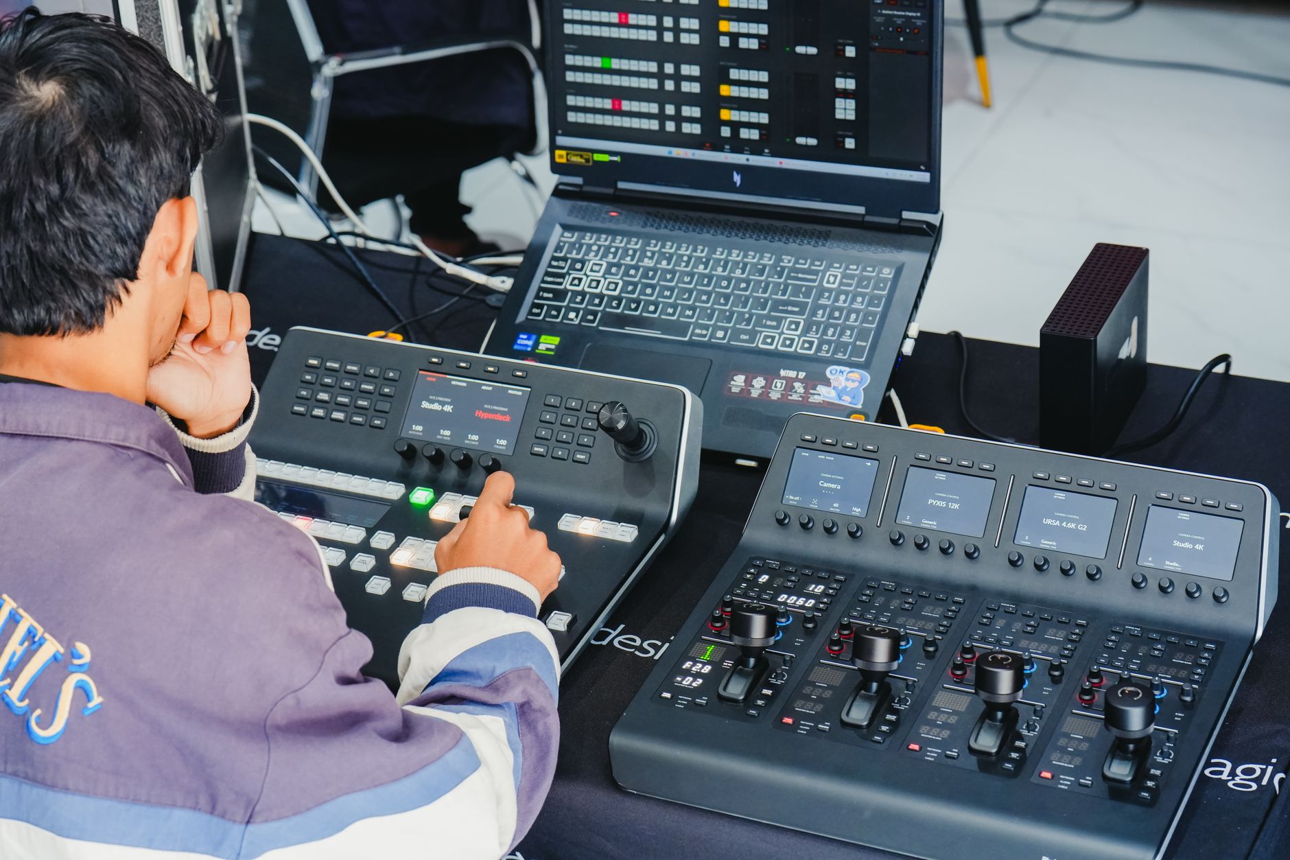 A person in a purple and white jacket operates a control panel, with a laptop and a second console with joysticks on a table.