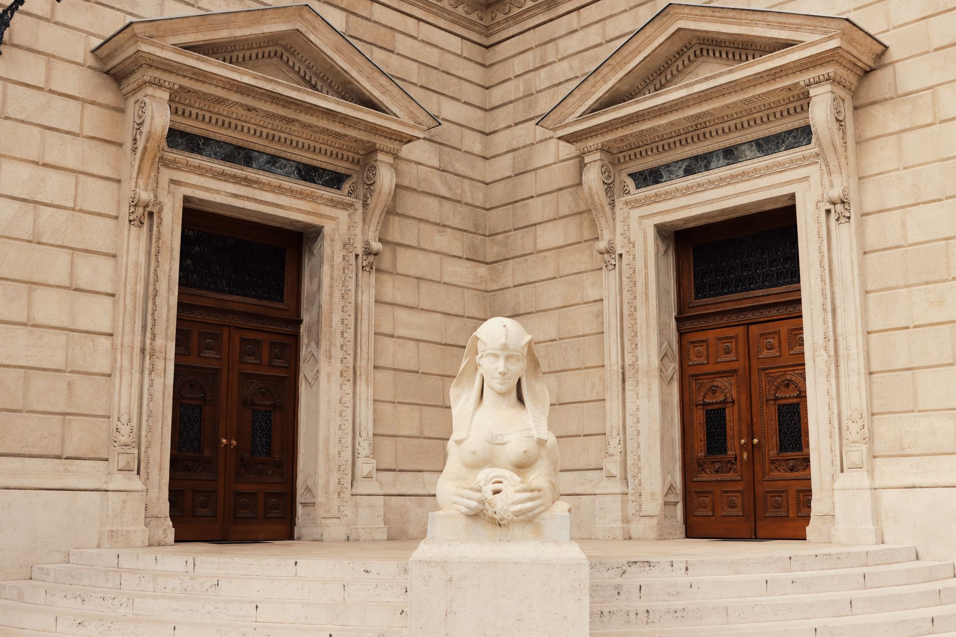 Stone building facade with two ornate carved doorways, wooden double doors and a white statue in an Egyptian-style headdress on a pedestal in front of steps.