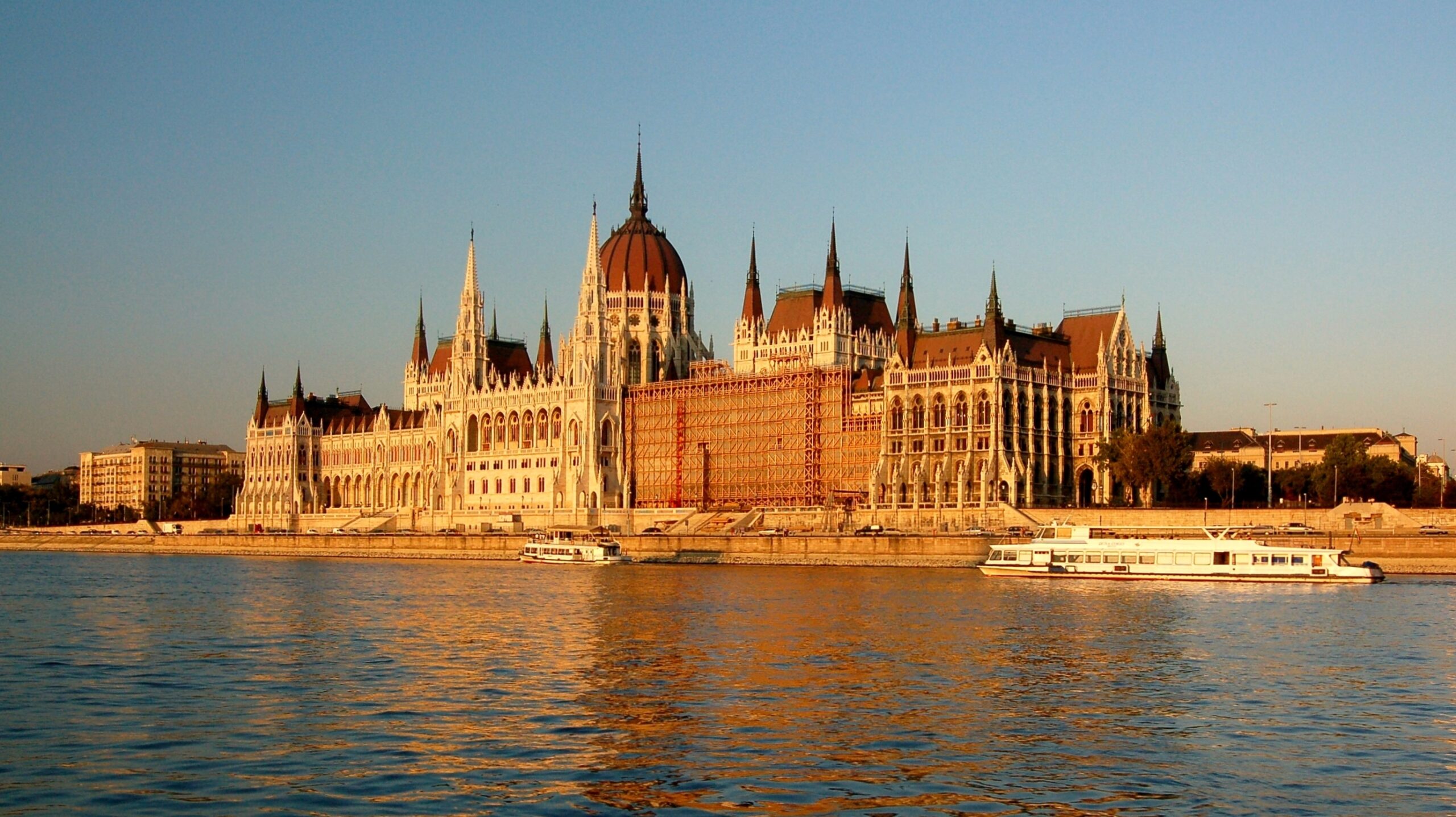 The Hungarian Parliament Building along the Danube riverfront in Budapest with passenger boats on the water.