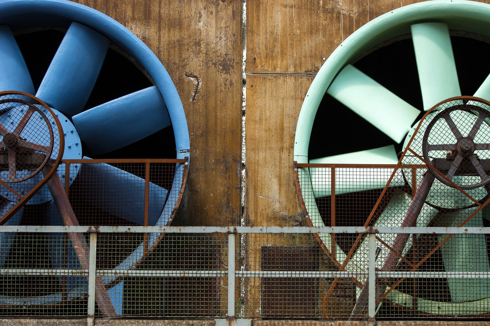 Two large industrial ventilation fans with blue and green rims installed on a weathered concrete wall behind a wire fence.