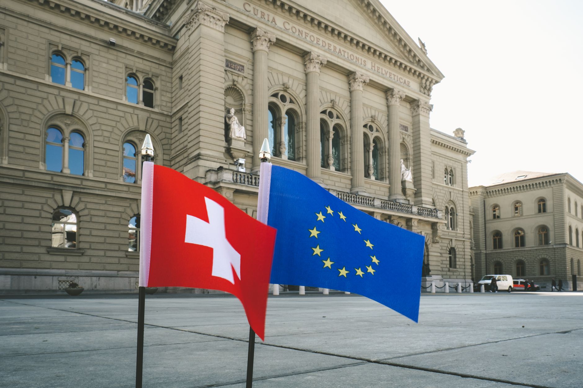 Un drapeau rouge à croix blanche et le drapeau européen devant un grand bâtiment parlementaire en pierre, sur une place dallée.