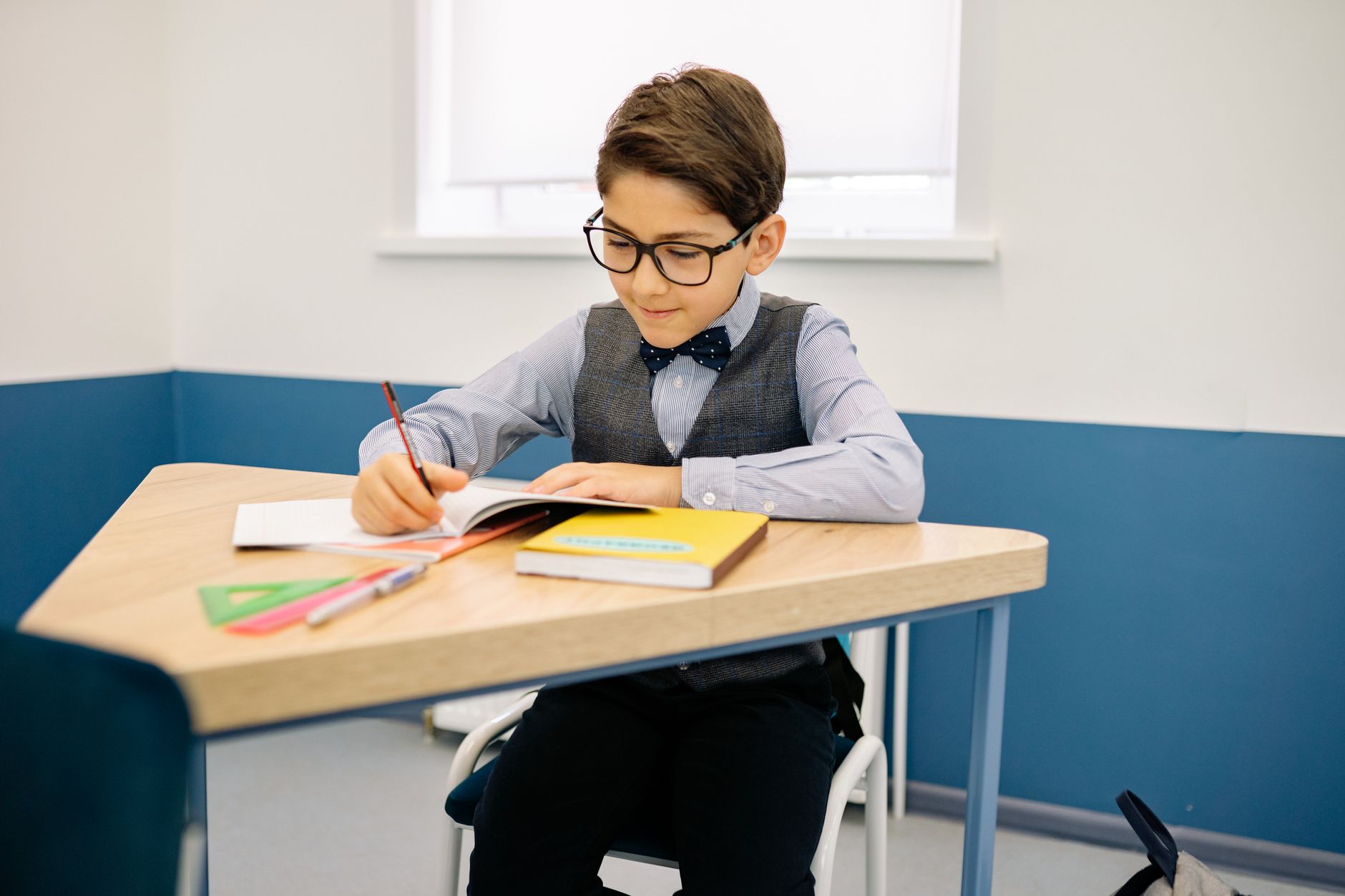 Un garçon à lunettes et nœud papillon écrit dans un cahier à son pupitre, dans une salle de classe, avec un livre jaune et une équerre verte posés devant lui.